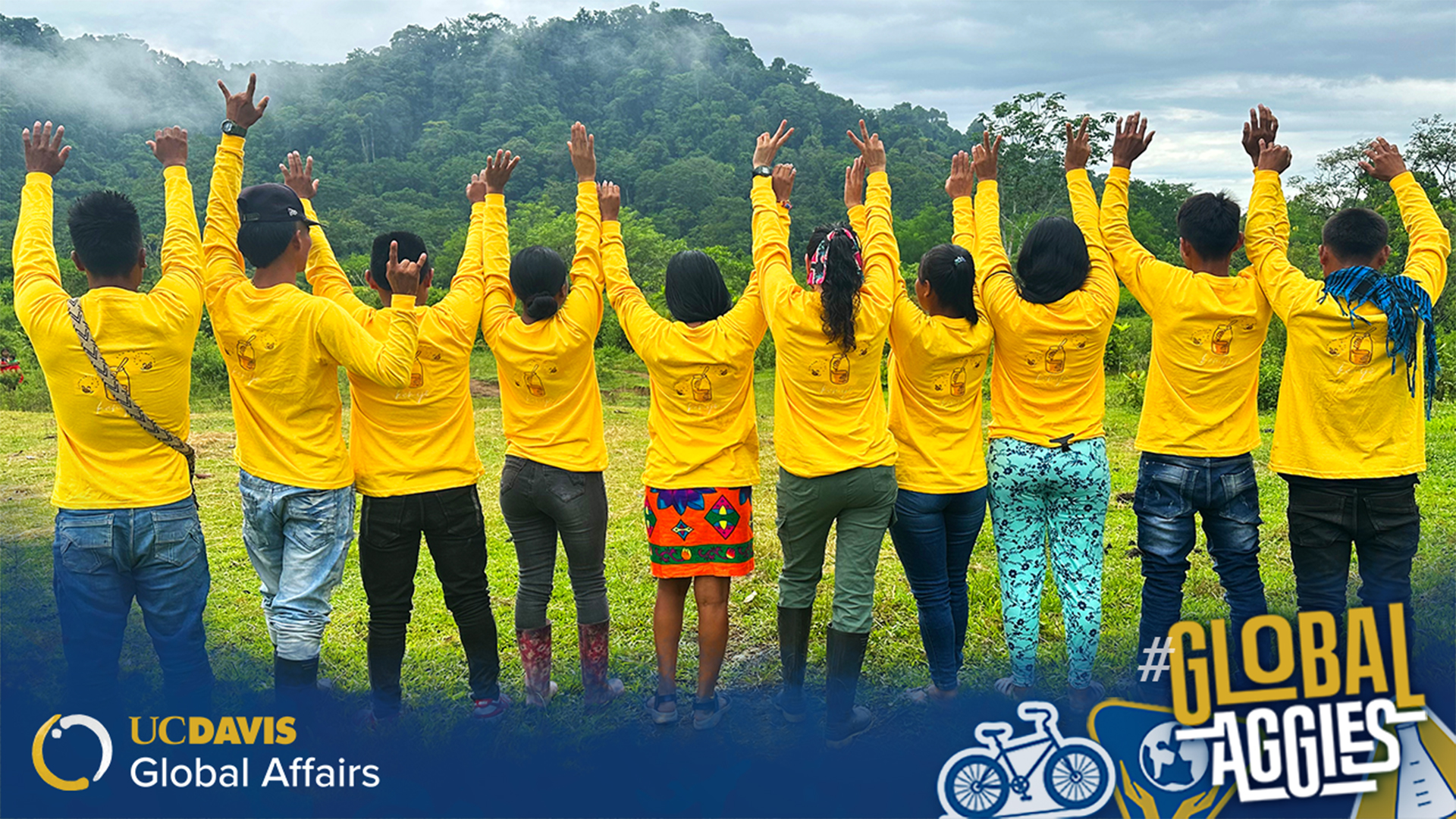 Group of ten people standing outdoors with their backs to the camera, all wearing bright yellow long-sleeve shirts and raising their arms in celebration. They are surrounded by lush green hills and trees under a cloudy sky. The bottom of the image displays the UC Davis Global Affairs logo and the hashtag '#GlobalAggies' with icons of a bicycle and a globe.