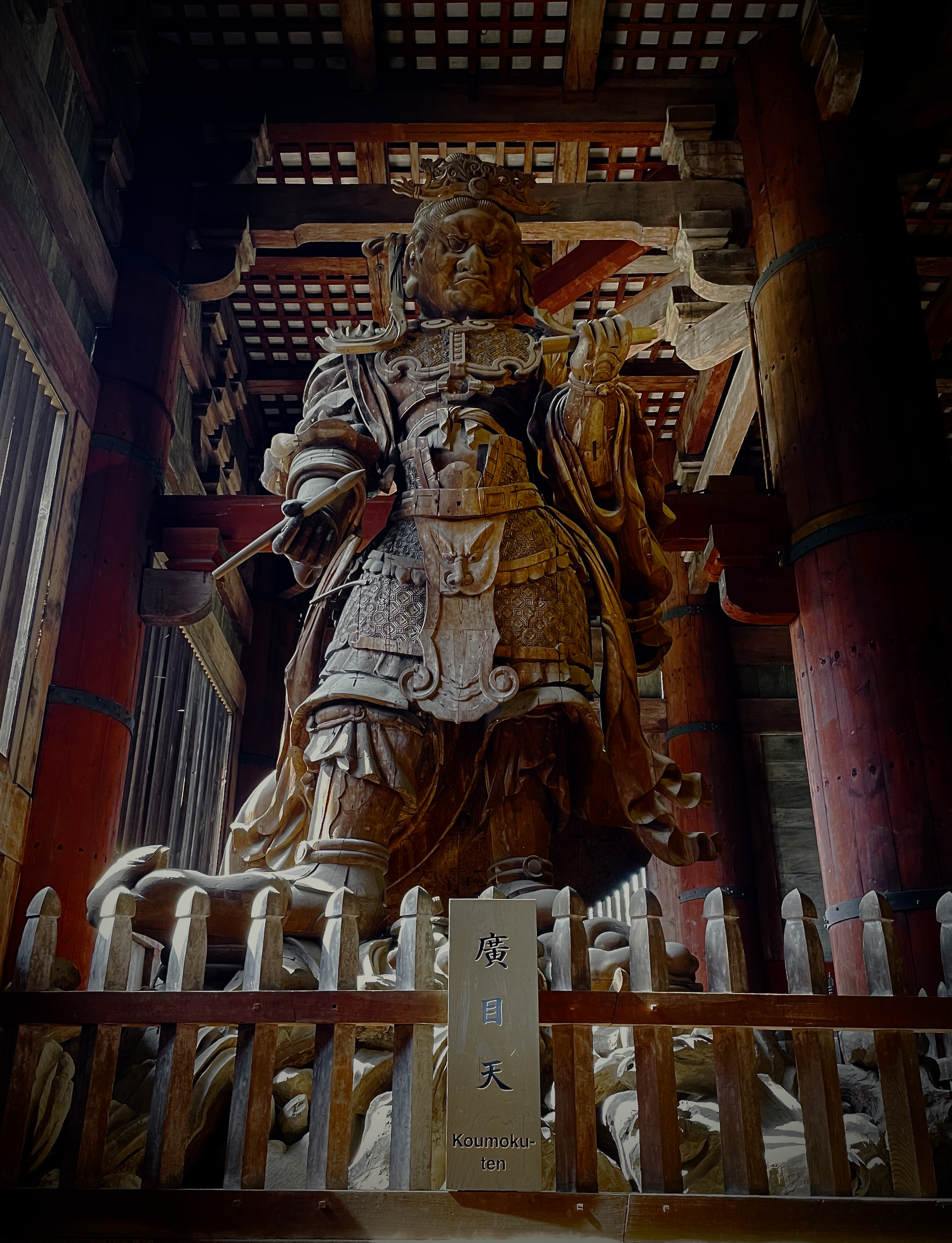 A large wooden statue of Koumokuten, an armored guardian deity, stands inside a historic temple surrounded by wooden beams and soft natural light.