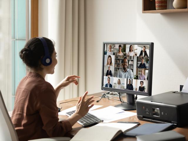 A person wearing headphones sits at a desk participating in a video conference call with multiple people displayed on a computer screen.