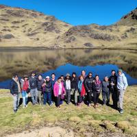 A group of people standing by a calm lake with grassy hills and a clear blue sky in the background.