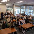 A diverse group of students smiling in a classroom setting with tables and large windows.