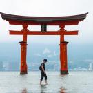 A man stands in calm water, reflecting the serene surroundings and the clear sky above.