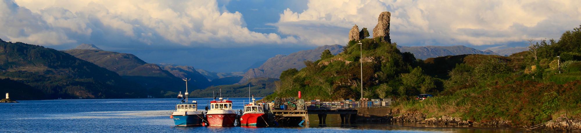 Small, colorful fishing boats docked at a pier on a calm blue lake, with a grassy hillside and ancient stone ruins in the background under a sky filled with fluffy white clouds.
