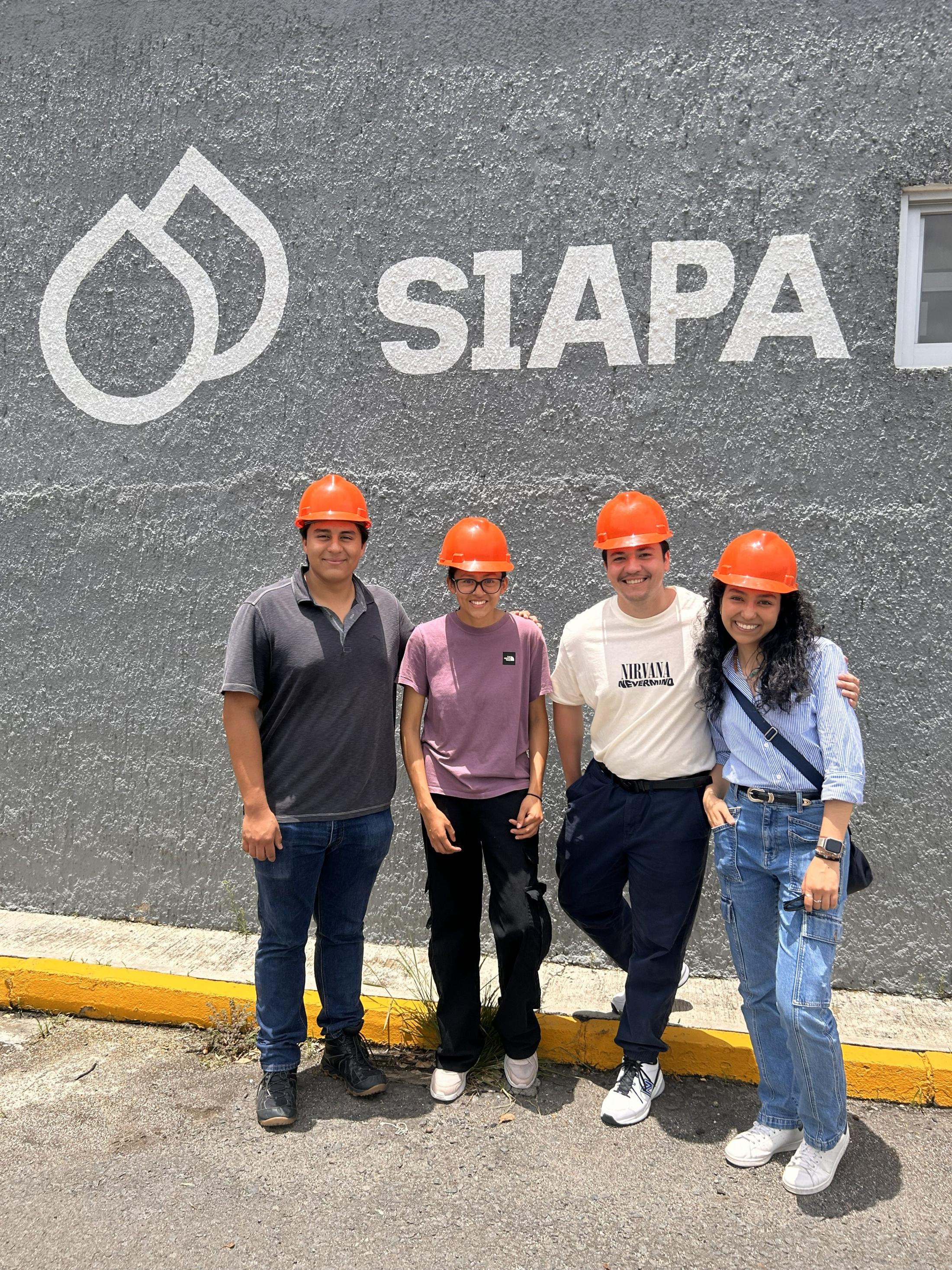 Four people in orange hard hats stand in front of a gray wall with "SIAPA" written on it.