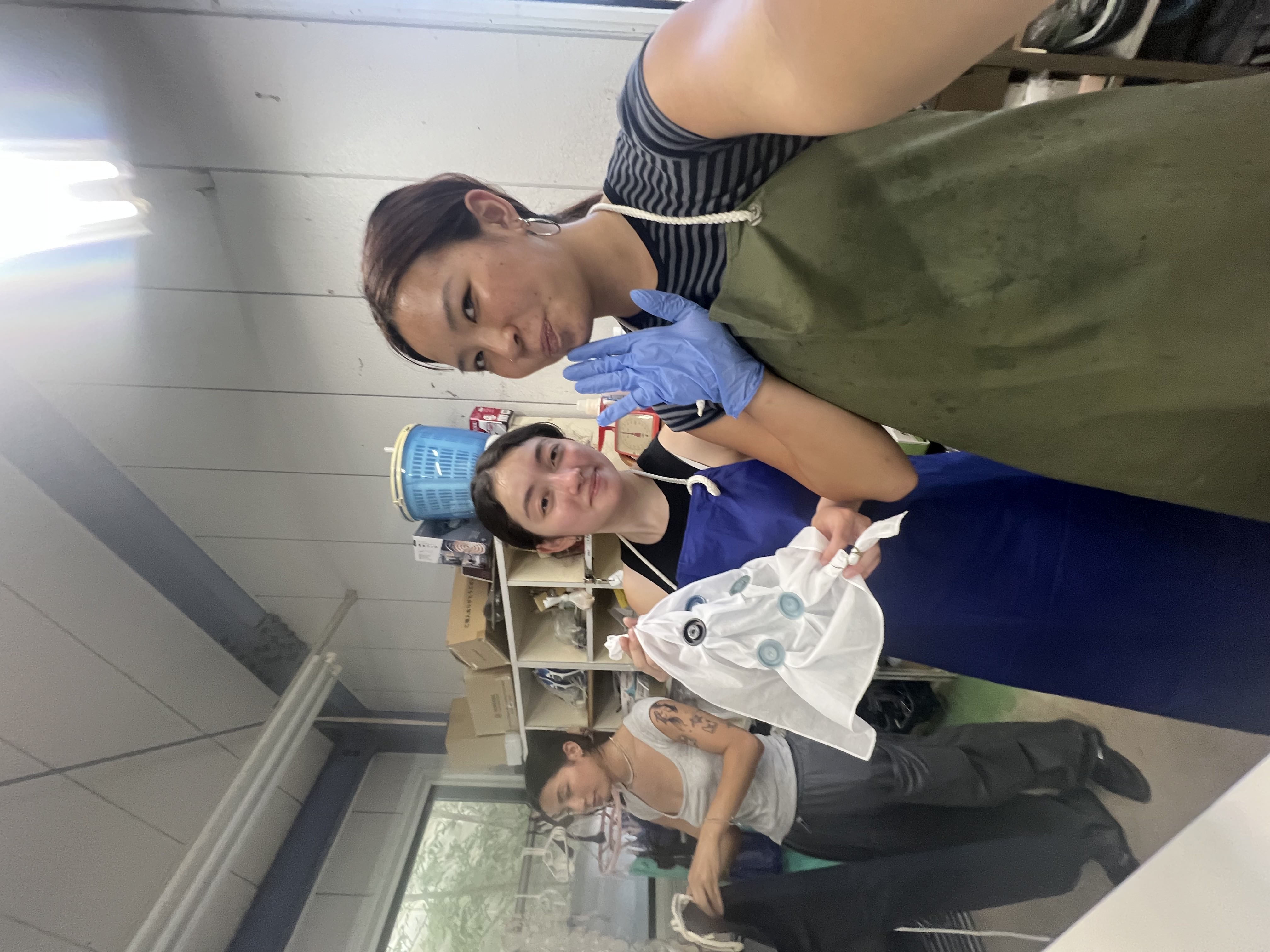 Two women smiling in a workshop, one wearing gloves and holding a towel.