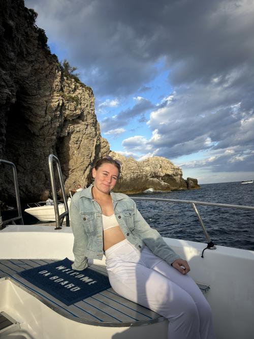 Young woman seated on boat bow by rocky cliff, denim jacket, cloudy sky (photo)