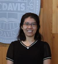 Smiling woman with glasses wearing black top standing before wooden wall and circular emblem