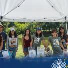 Seven smiling volunteers at outdoor booth under white canopy by lake; Global Aggies logo