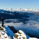 A mountaineer stands on a snow-covered mountain peak, taking a photo of a vast landscape of snowy mountains, clouds, and a lake below under a clear blue sky.