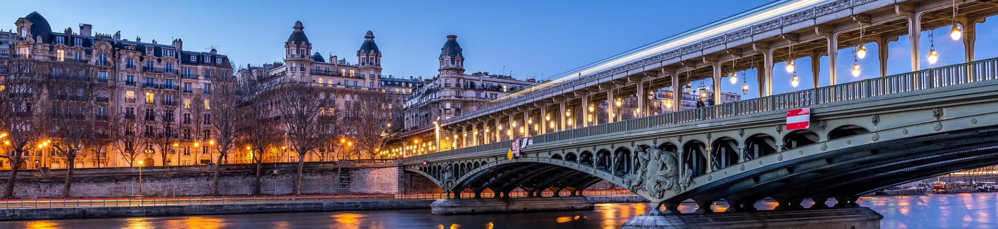 A bridge illuminated at night, reflecting in the calm river under a blue sky.